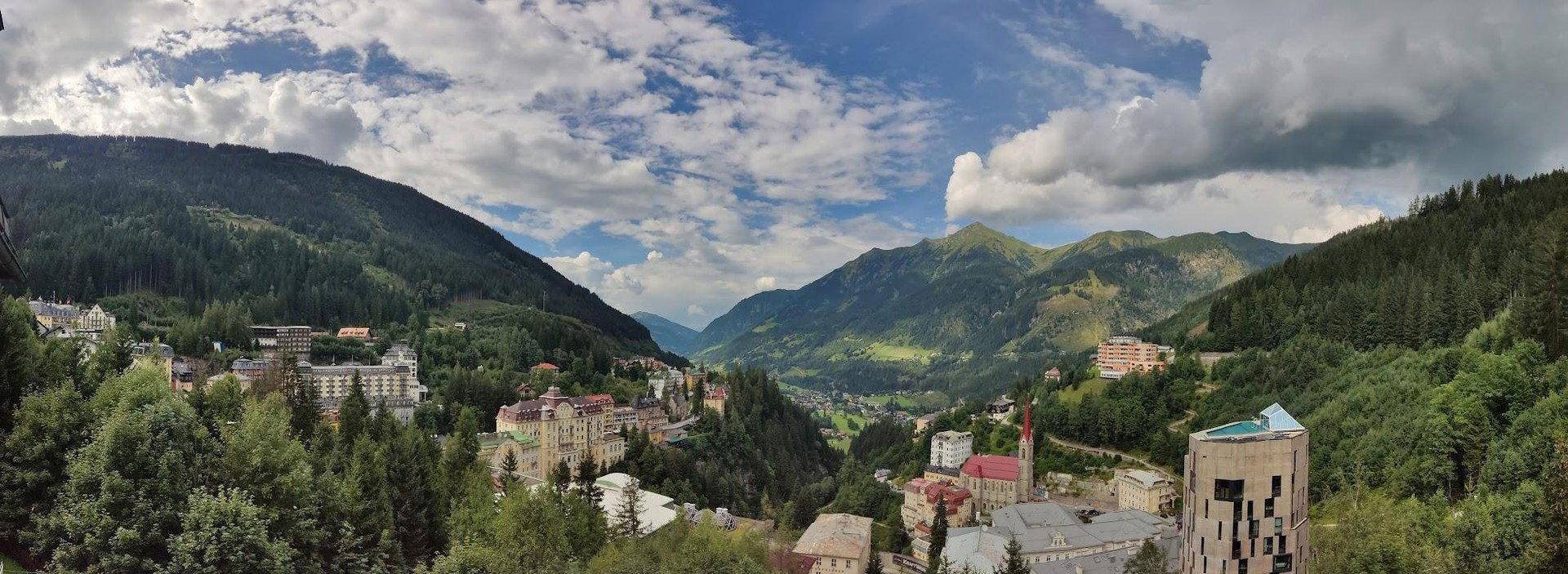 Weitläufiger Panoramablick auf ein grünes Tal mit einem Dorf, umgeben von dichten Wäldern und Bergen unter blauem Himmel.