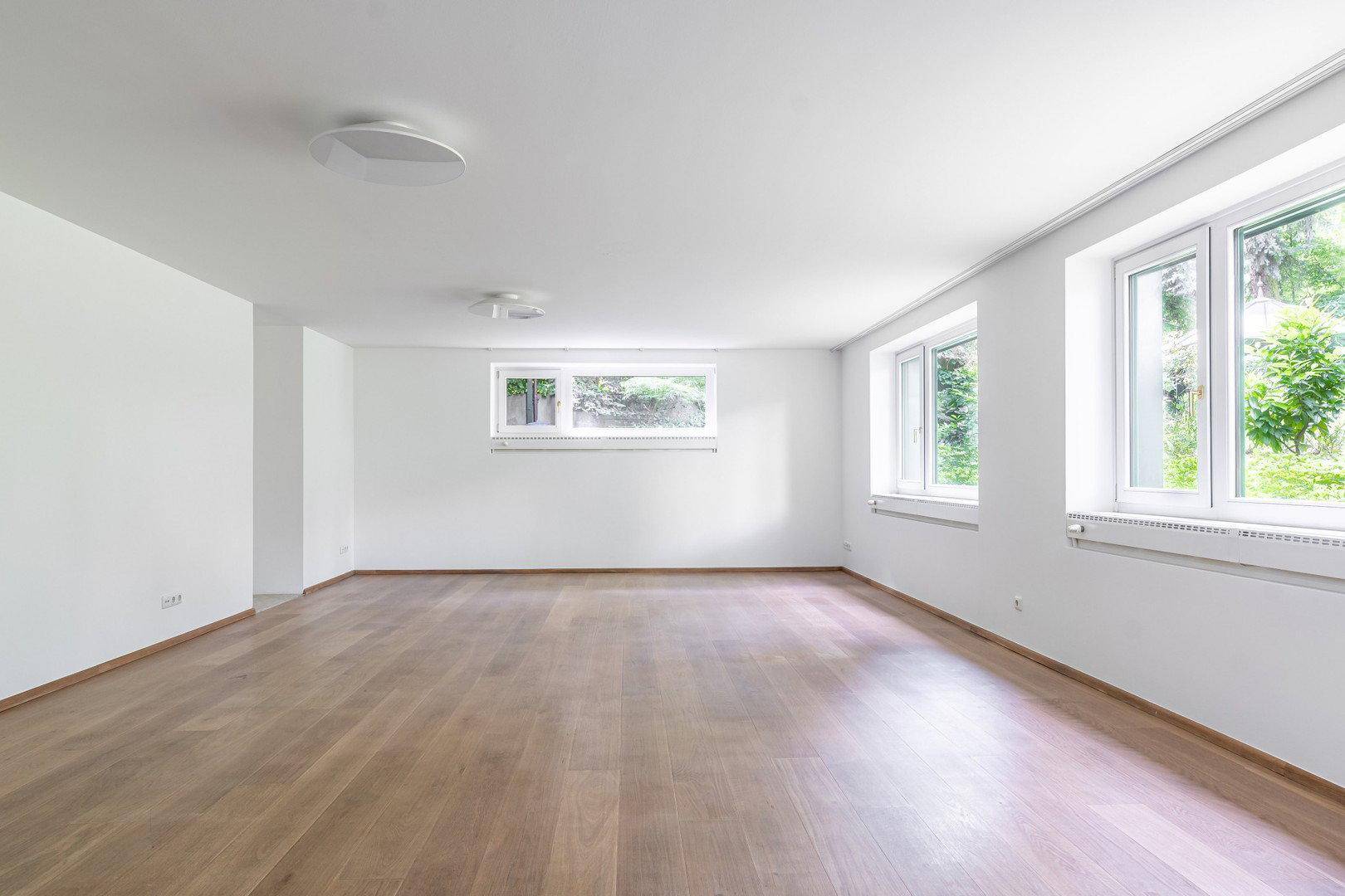Geräumiger, leerer Raum mit hellem Holzboden und großen Fenstern zum Garten.