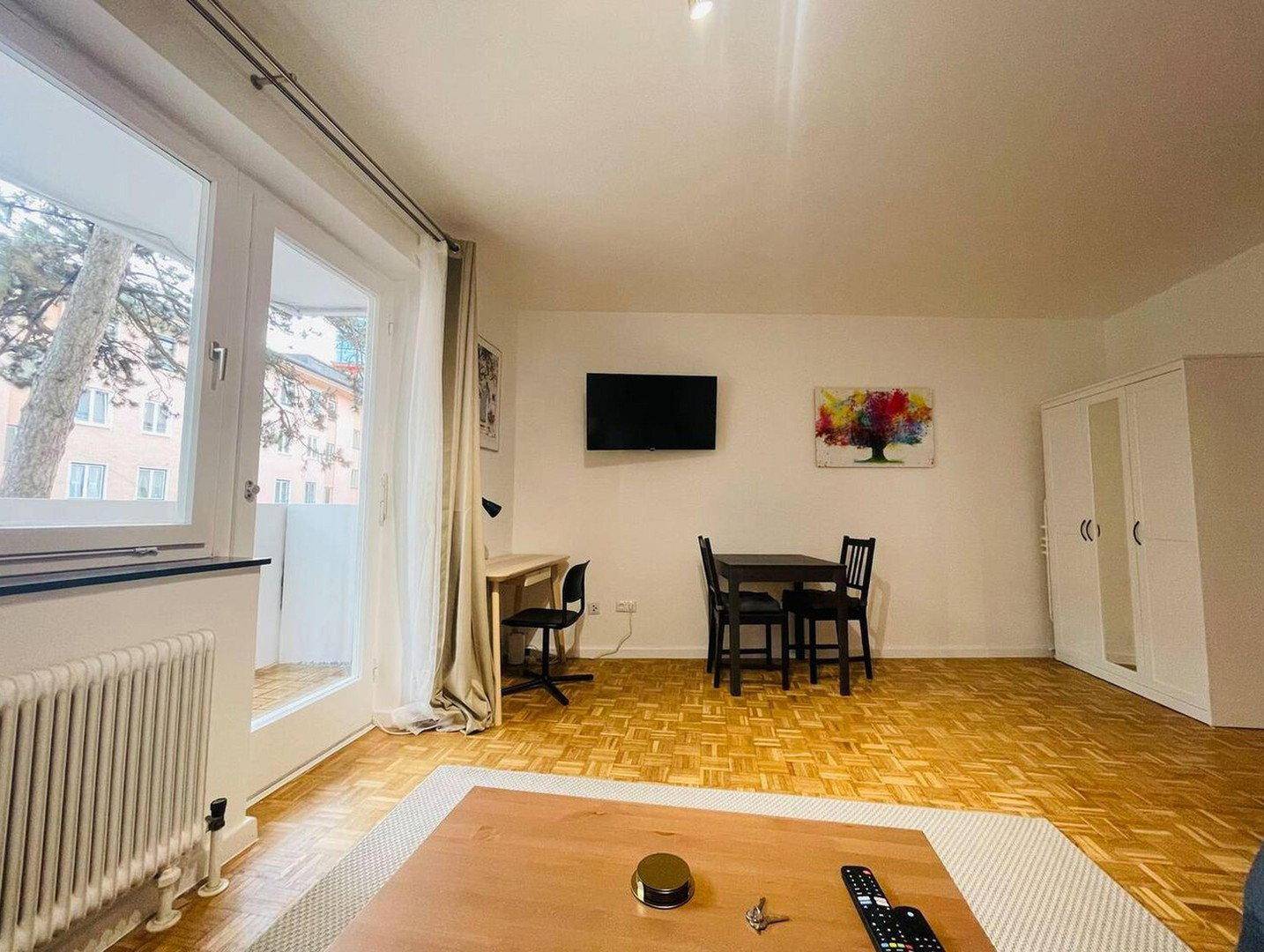 Heller Wohnbereich mit Parkettboden, Fernseher, Schreibtisch und Zugang zum Balkon.