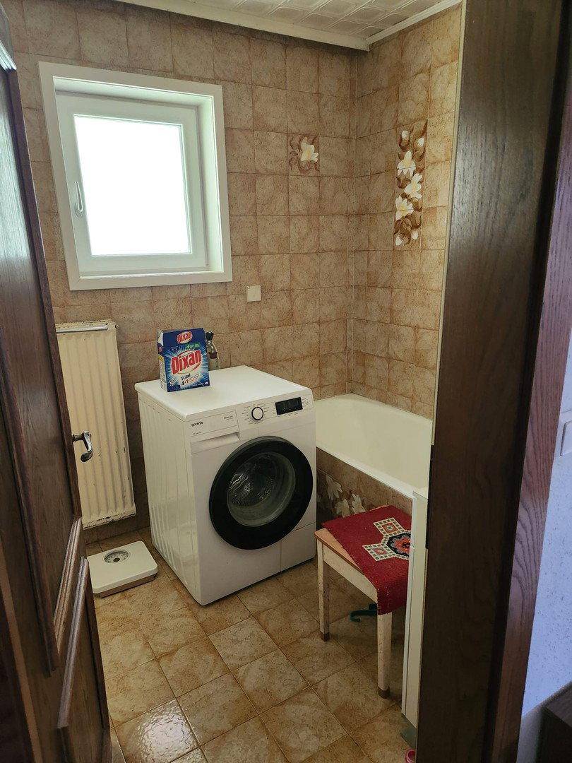 Badezimmer mit gefliesten Wänden und Boden, einer Badewanne, Waschmaschine und einem Fenster.