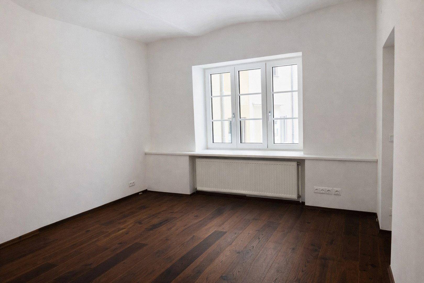 Helles Schlafzimmer mit dunklem Parkettboden und einem großen Fenster mit Blick ins Grüne.