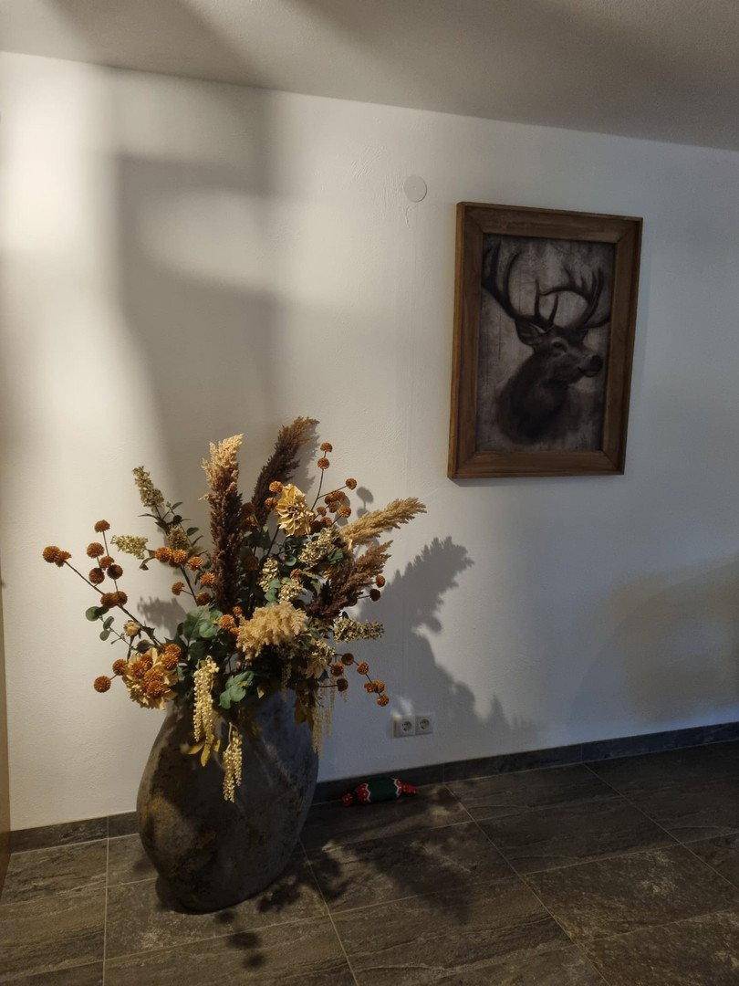 Dekorativer Bereich mit großer Vase, Trockenblumen und Hirschmotiv an der Wand.