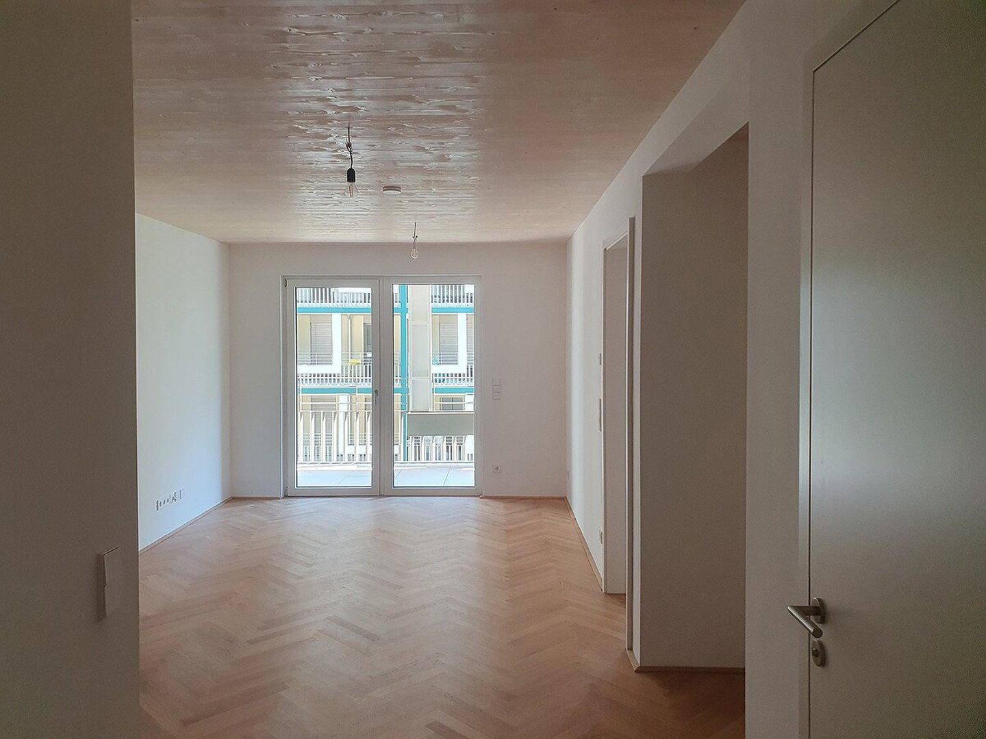 Heller, leerer Raum mit Fischgrätparkettboden, weißen Wänden und Zugang zu einem Balkon.