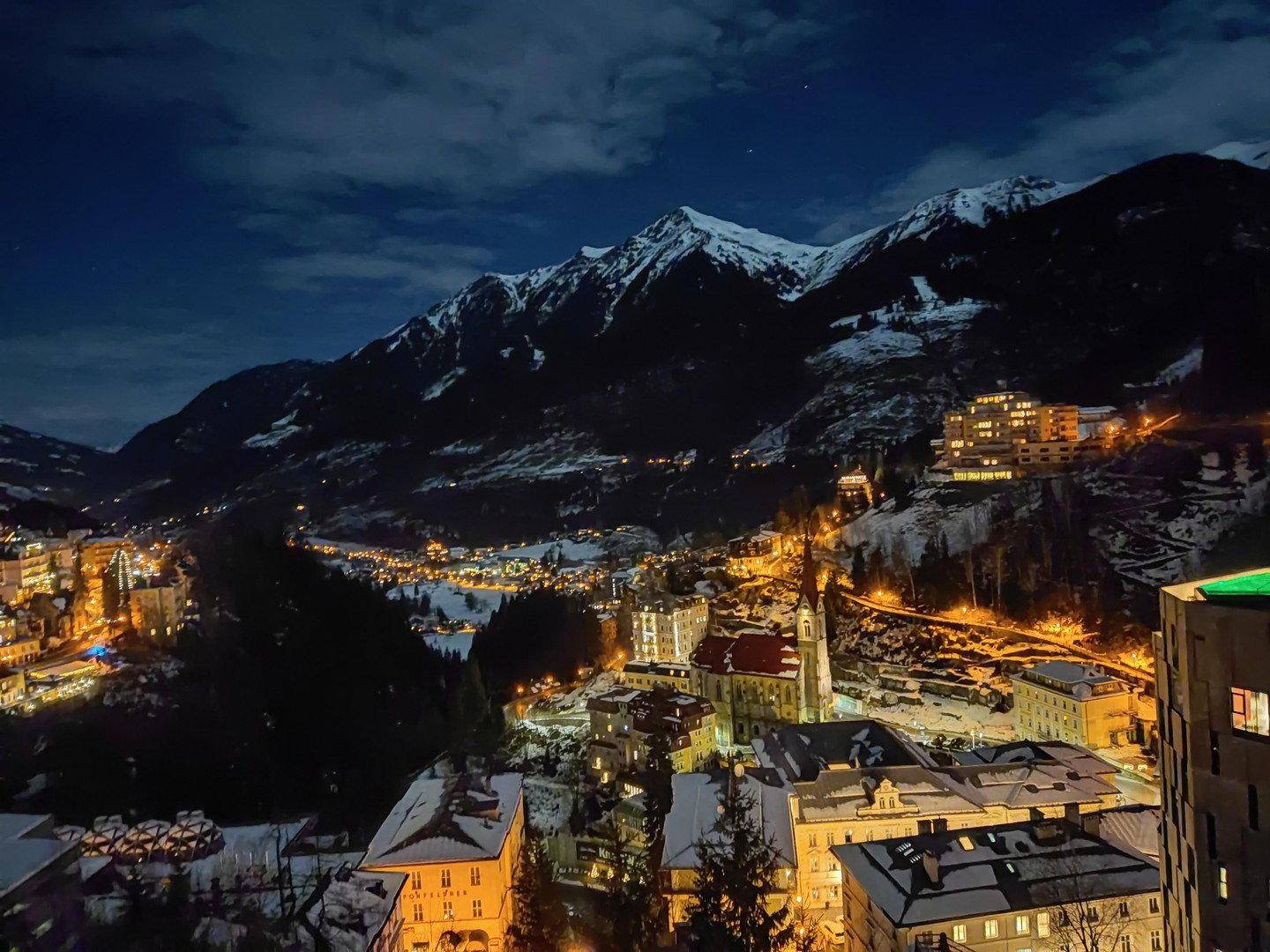 Beeindruckender Nachtblick auf ein beleuchtetes Dorf im Tal, umgeben von schneebedeckten Bergen unter klarem Sternenhimmel.