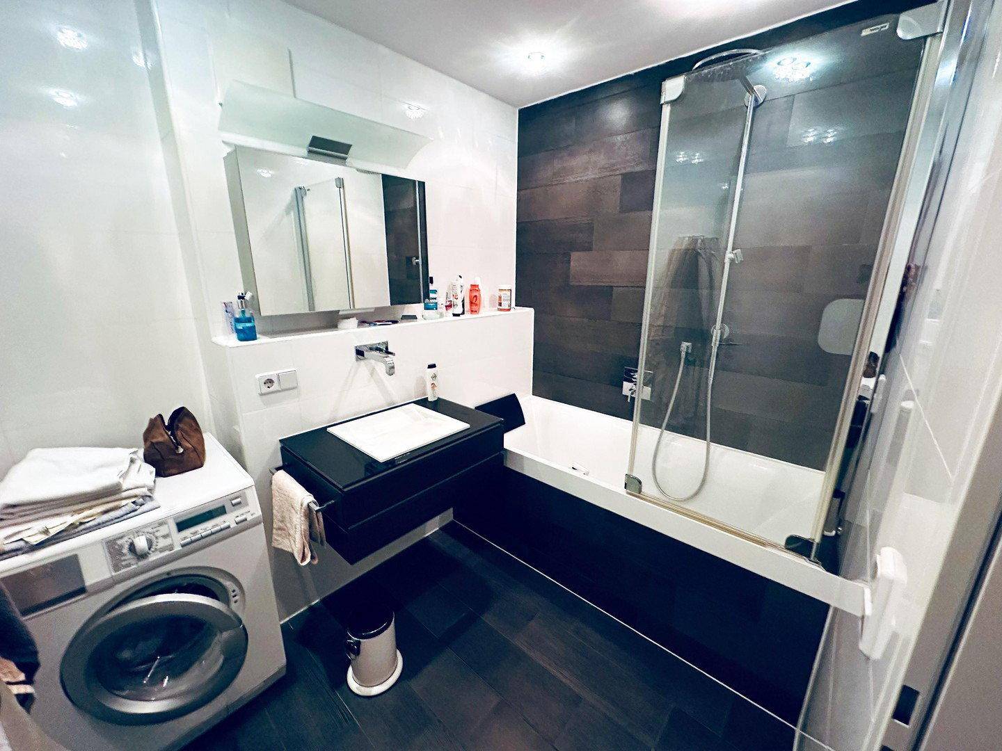 Modernes Badezimmer mit Badewanne, Dusche, Waschmaschine und dunklen Fliesen.