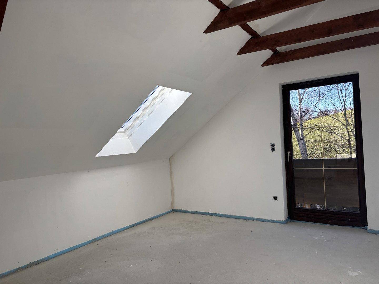 Dachbodenraum mit Dachfenster und Tür, die einen Blick ins Grüne ermöglicht, im Rohbauzustand.