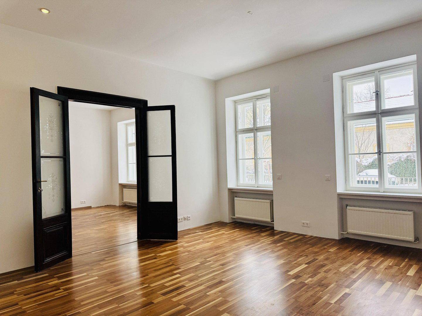 Geräumiges Wohnzimmer mit eleganten Flügeltüren, Parkettboden und großen Fenstern.