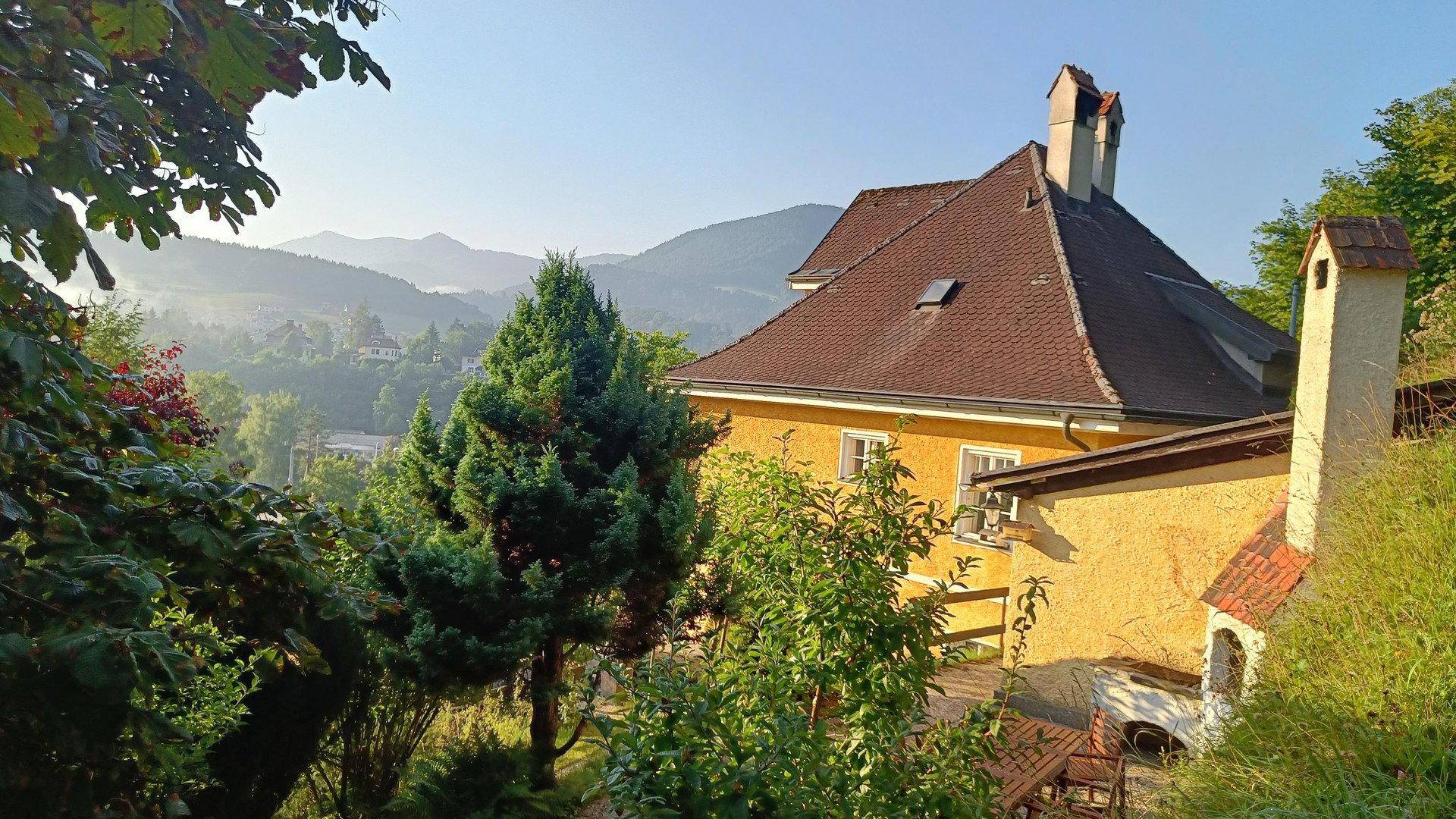 Historisches Gebäude mit markantem Dach und Schornstein, eingebettet in eine hügelige Landschaft.