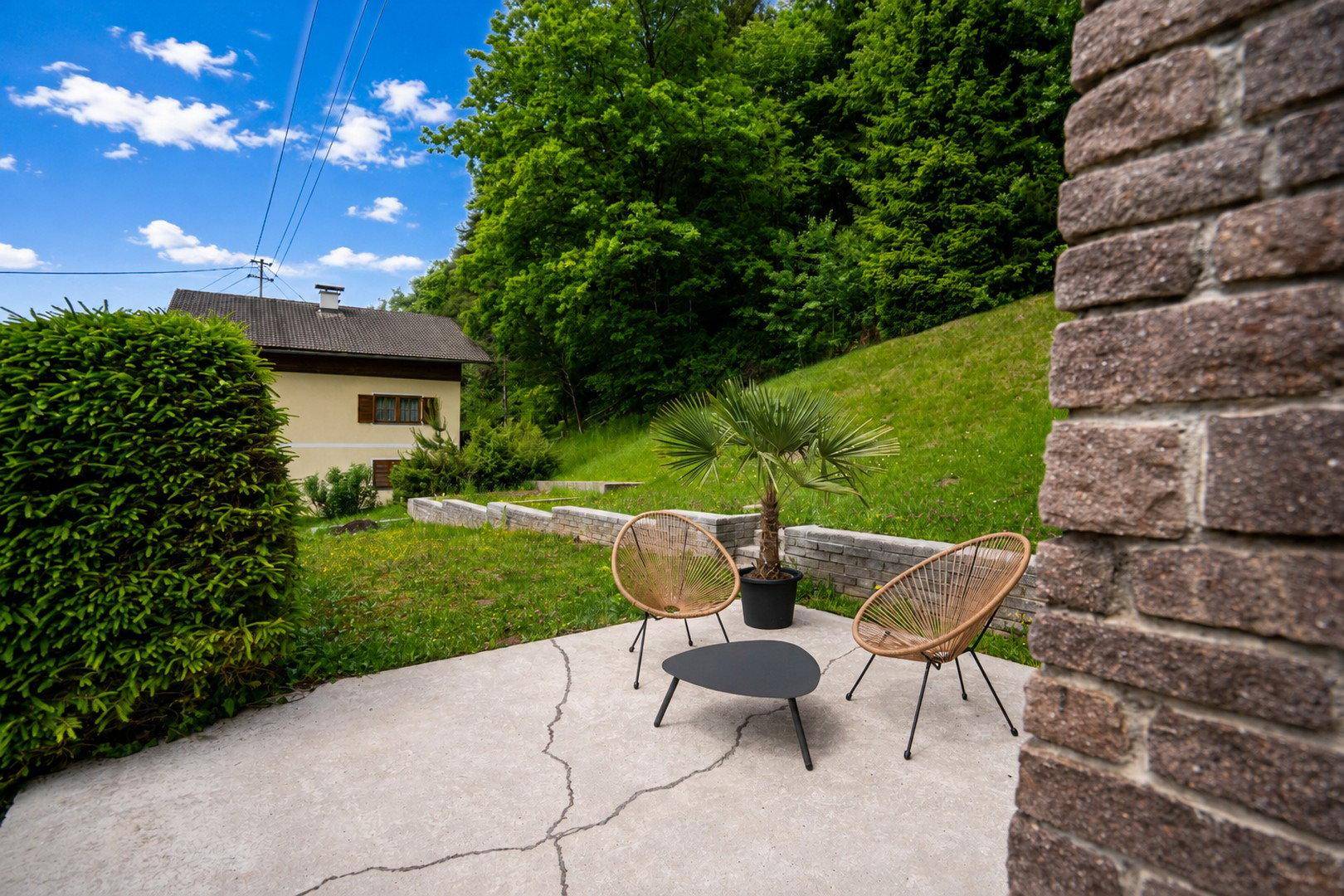 Gemütliche Terrasse mit Korbmöbeln, Palmen und einer Ziegelwand im Grünen.