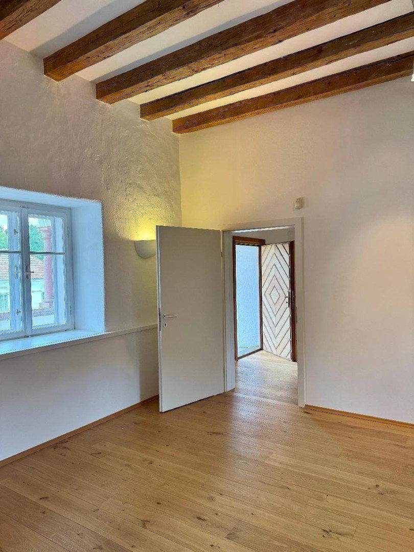 Heller Raum mit Holzbalken, Holzboden und einem Fenster mit Blick nach draußen.