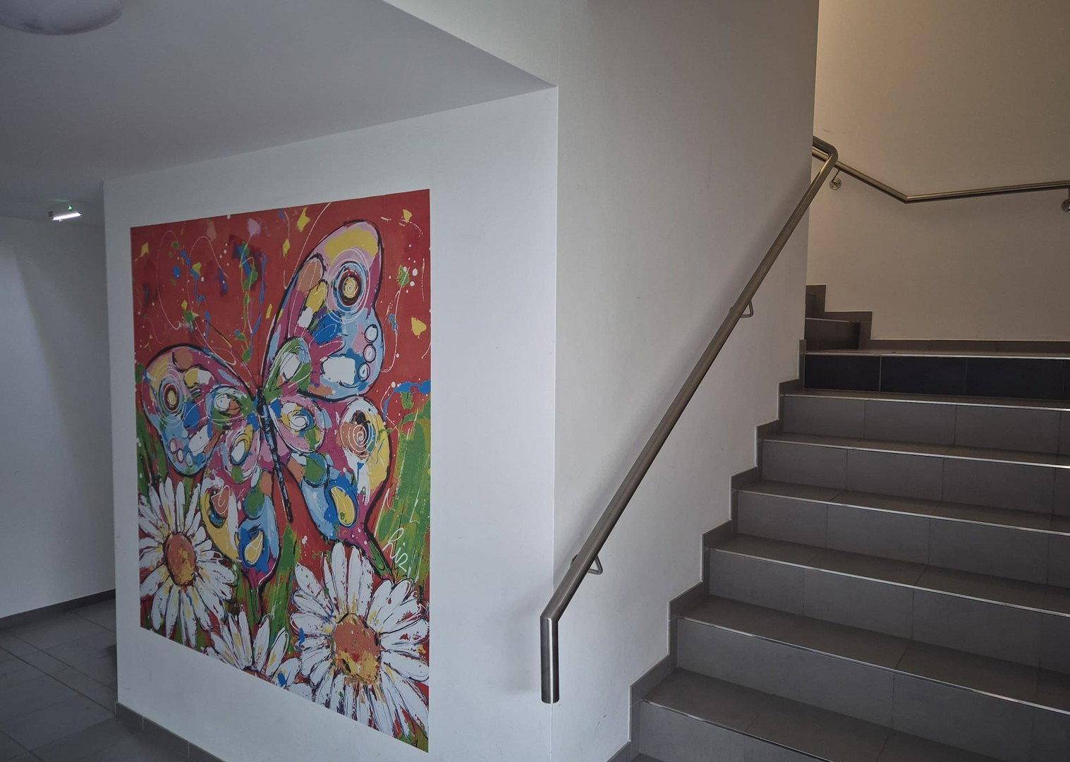 Helles Treppenhaus mit modernem Geländer und farbenfrohem Wandbild.