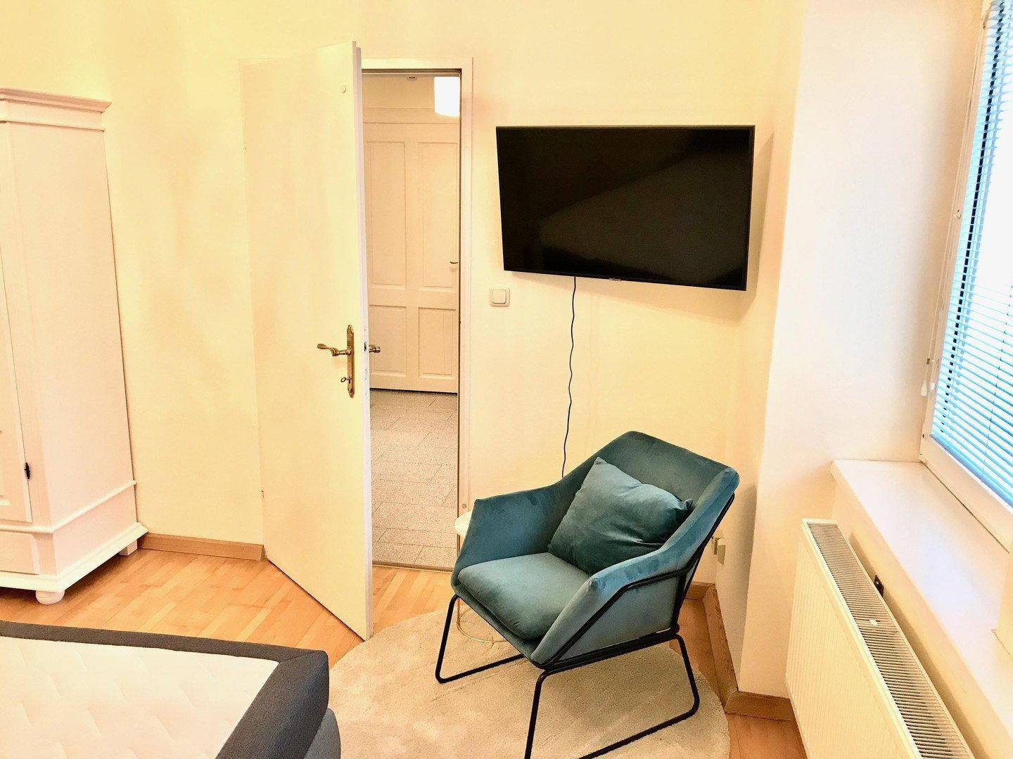 Ecke eines Schlafzimmers mit bequemem Sessel, Wandfernseher und Zugang zu einem weiteren Raum.