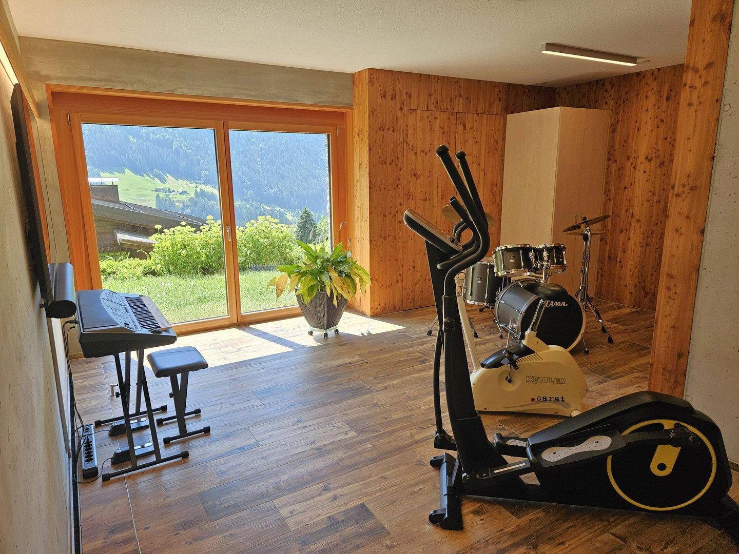 Fitnessraum mit Trainingsgeräten, Musikinstrumenten und großen Fenstern mit Bergblick.