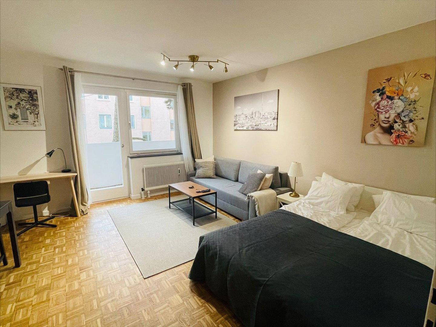 Heller Wohn- und Schlafbereich mit Schreibtisch, Sofa, Bett und Zugang zum Balkon.