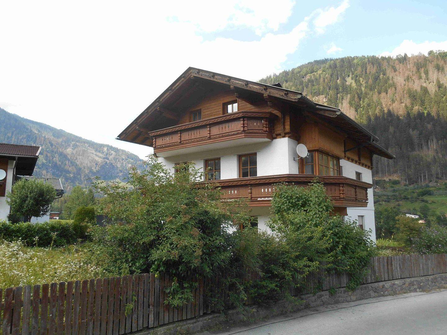 Traditionelles Haus mit Holzbalkonen und weißer Fassade, umgeben von üppiger Vegetation und Bergen.