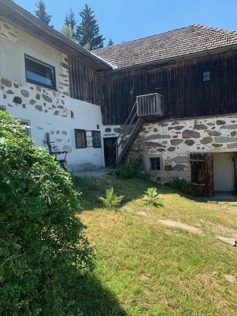 Historisches Gebäudeensemble mit Steinmauern, Holzfassaden und einer Außentreppe im Grünen.
