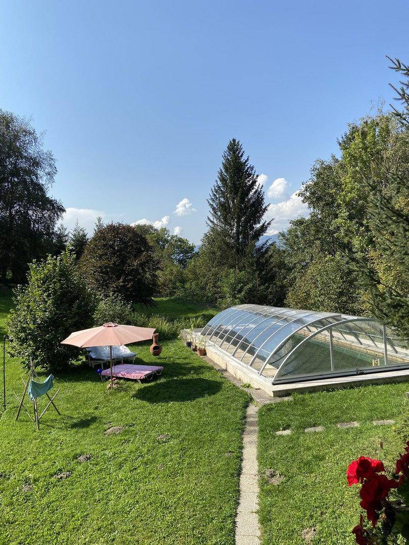 Sonniger Gartenbereich mit überdachtem Pool, Liegewiese und schattenspendenden Bäumen.