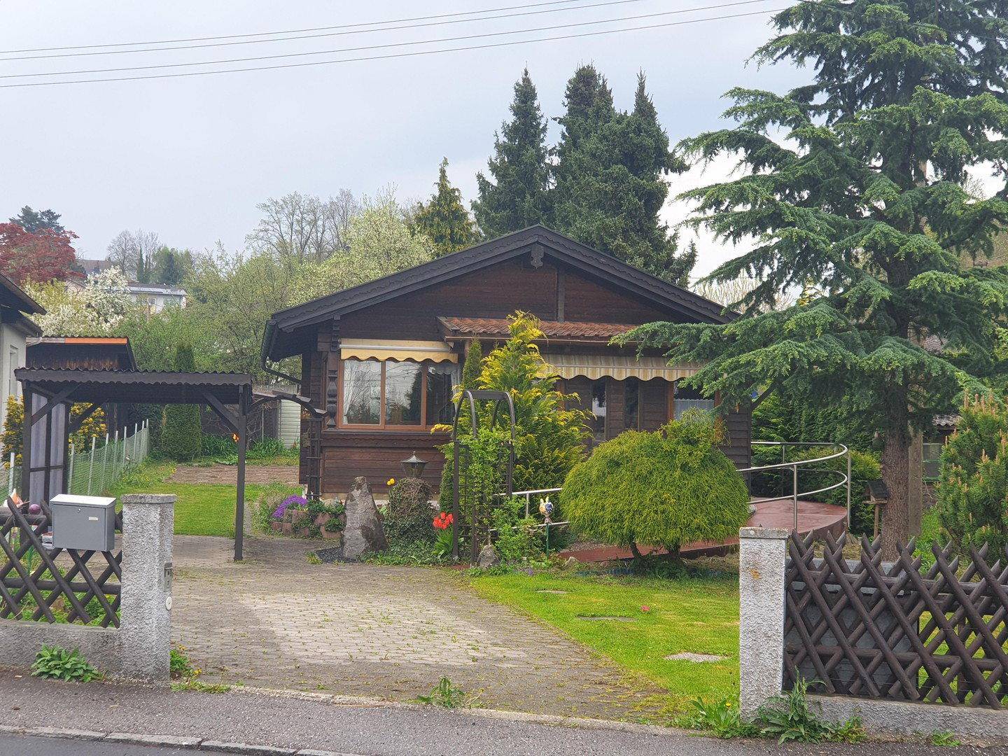 Ansicht des Holzhauses von der Straße mit Einfahrt, Carport und gepflegtem Vorgarten.