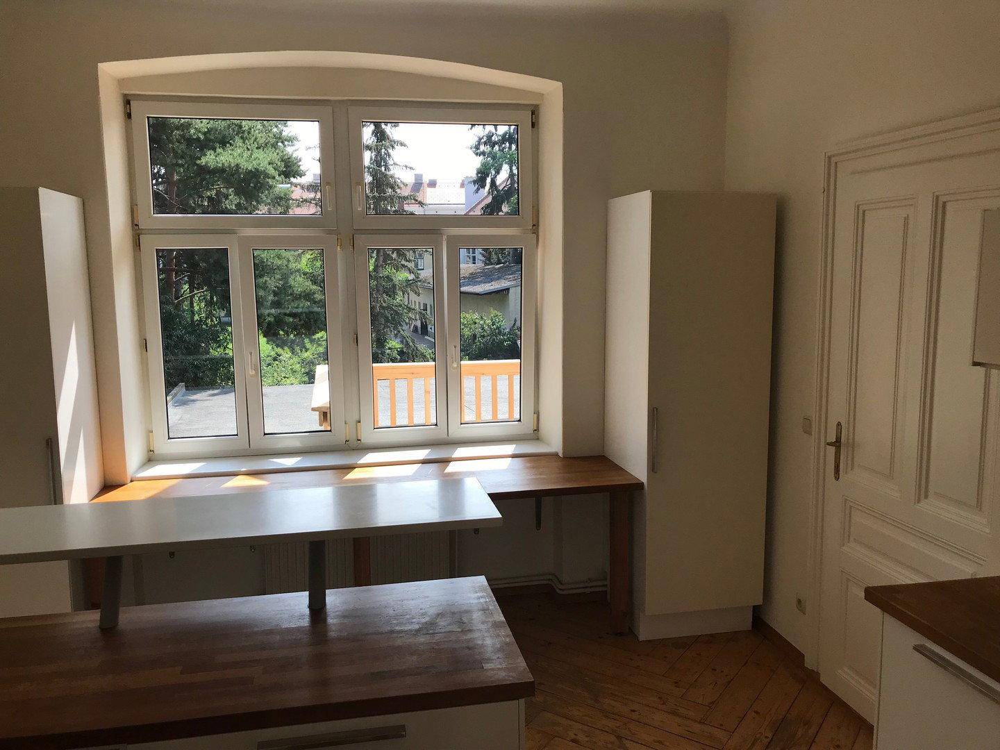 Heller Küchenbereich mit großem Fenster, Holztisch und weißen Schränken, die viel Tageslicht hereinlassen.