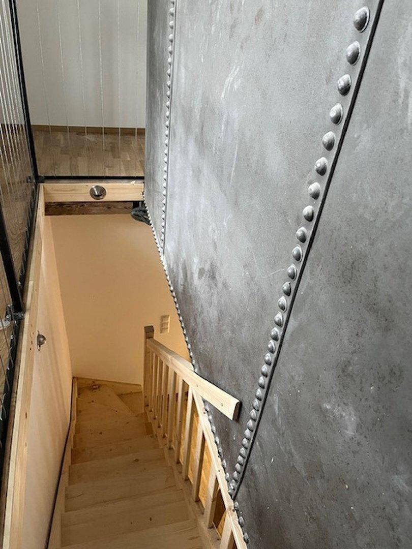 Treppenhaus mit Holzstufen und industrieller Metallwand, die nach unten führt.