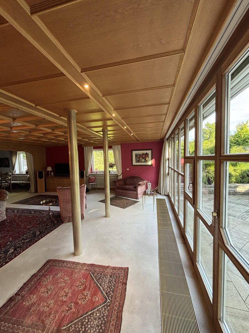 Großzügiges Wohnzimmer mit Holzkassettendecke, großen Fenstern und Blick auf die Terrasse.