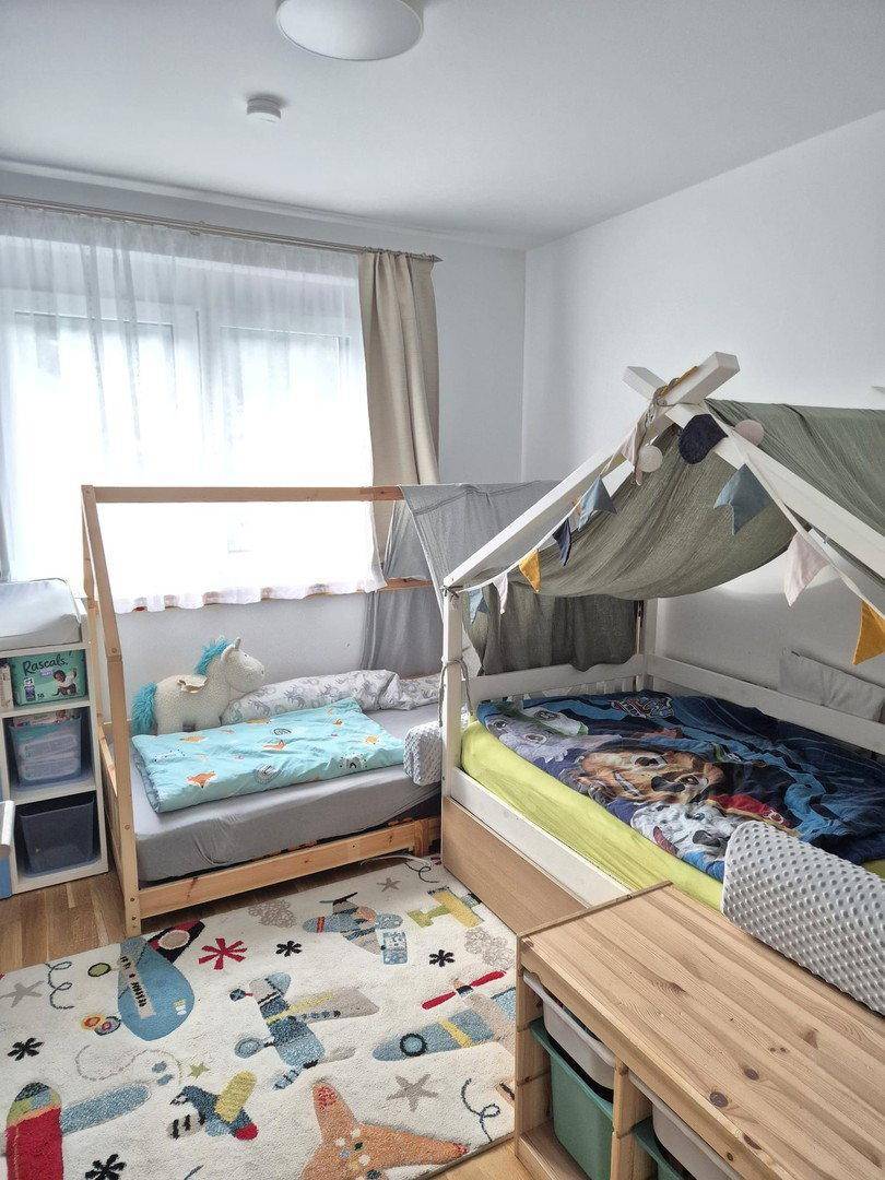 Helles Kinderzimmer mit zwei Betten, Spielteppich und viel Tageslicht.