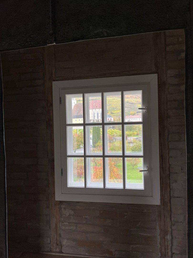 Kleines Fenster in einer rustikalen Wand mit Blick auf herbstliche Bäume und ein Dorf.