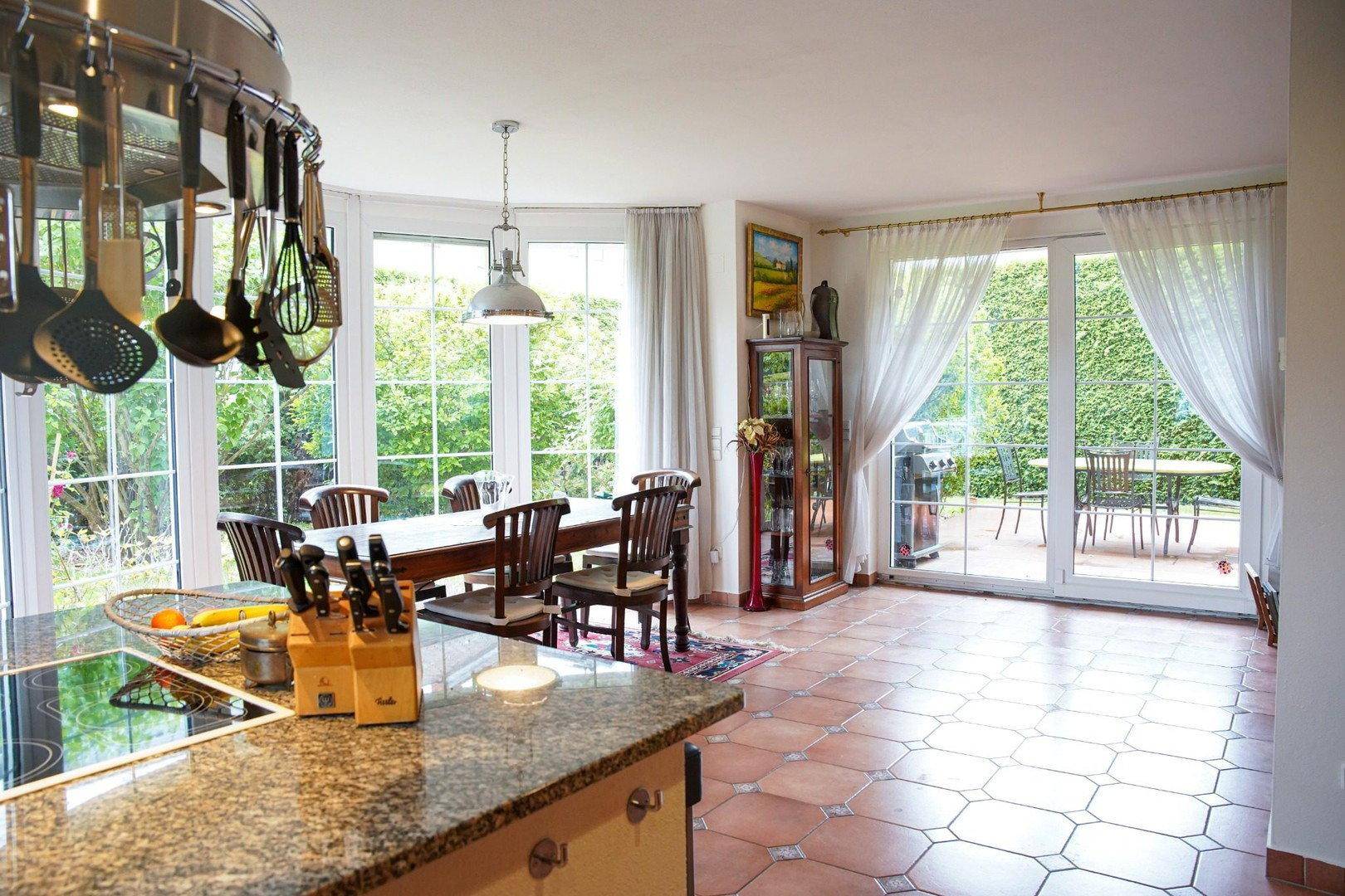 Offene Küche mit Essbereich und Zugang zur Terrasse, umgeben von großen Fenstern mit Gartenblick.