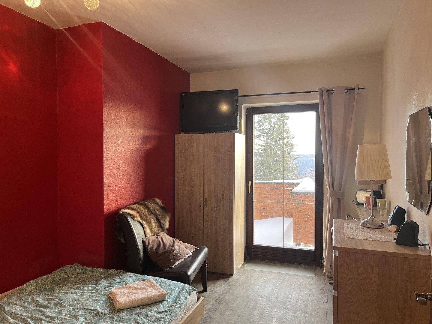 Schlafzimmer mit roter Akzentwand, Fernseher, Kleiderschrank und Zugang zu einem Balkon mit Winterblick.