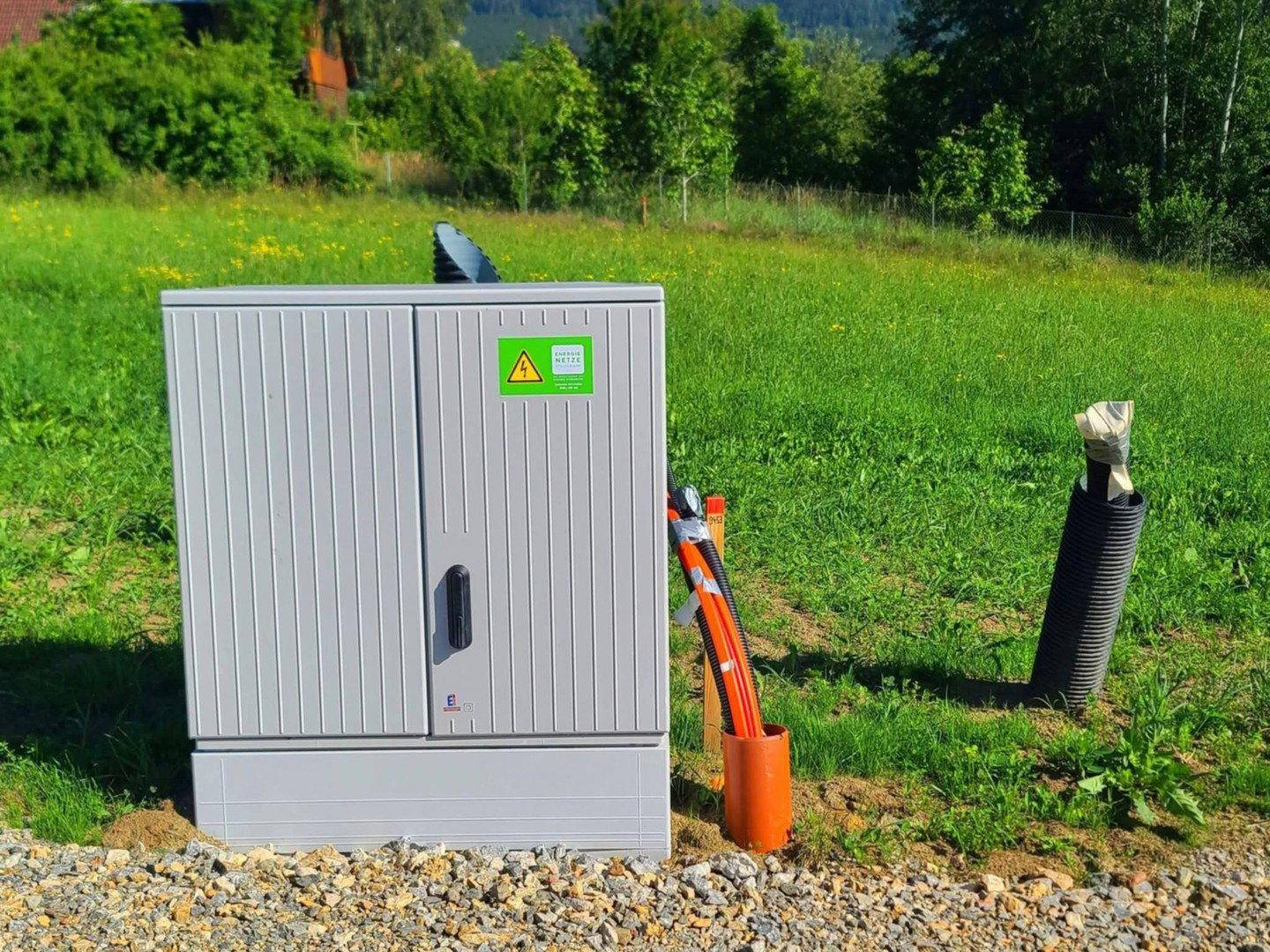 Grauer Versorgungskasten mit orangefarbenen Kabeln am Rande einer grünen Wiese.