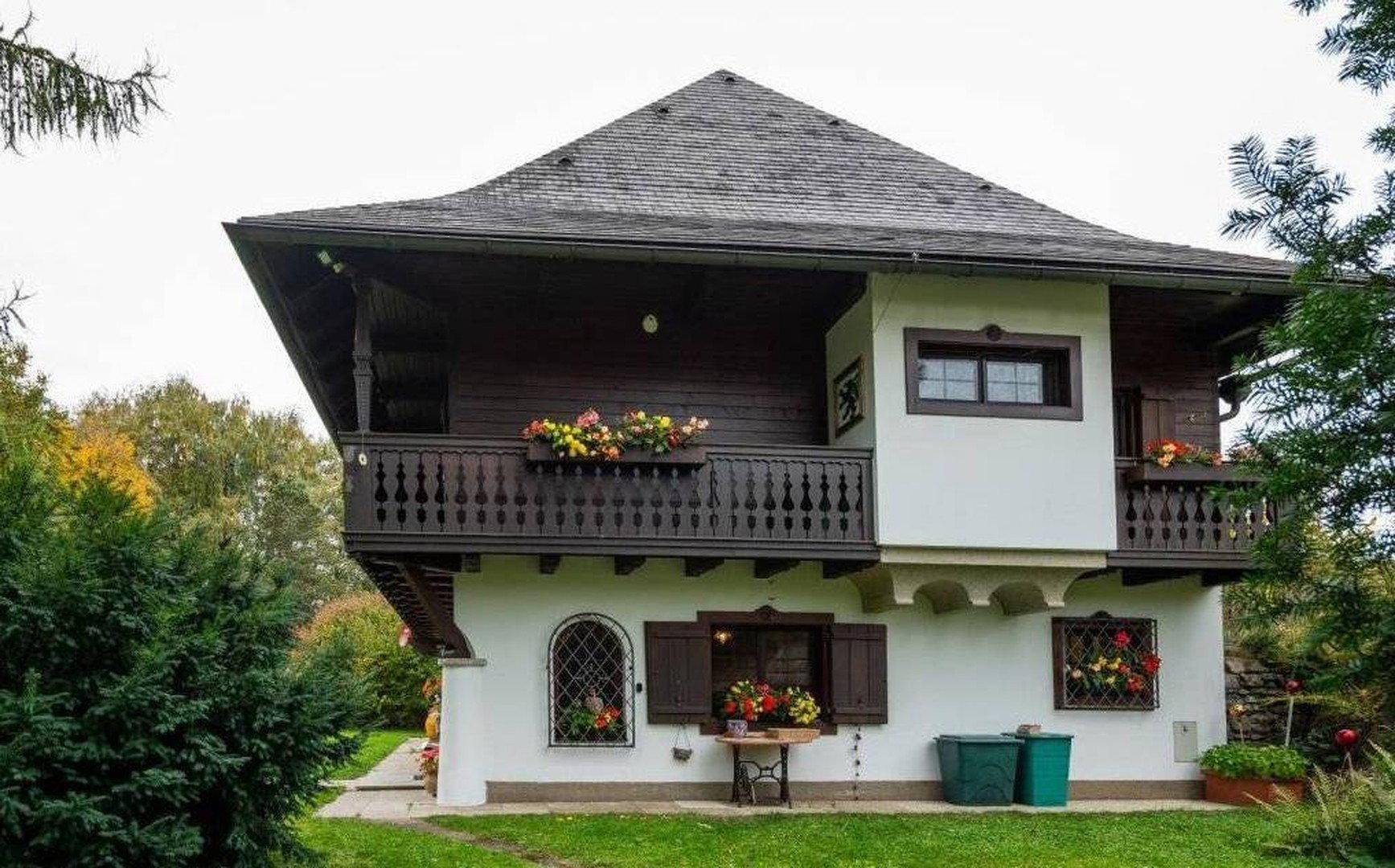 Seitliche Ansicht eines traditionellen Hauses mit Holzbalkonen, Blumenkästen und grüner Umgebung.