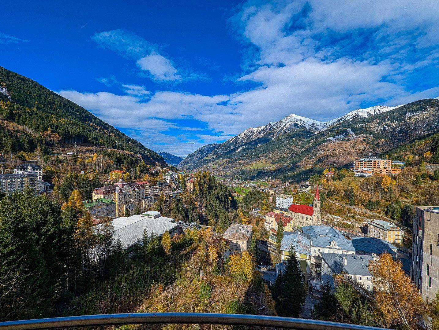 Panoramablick auf ein malerisches Tal mit einem Dorf, umgeben von bewaldeten Bergen und schneebedeckten Gipfeln.
