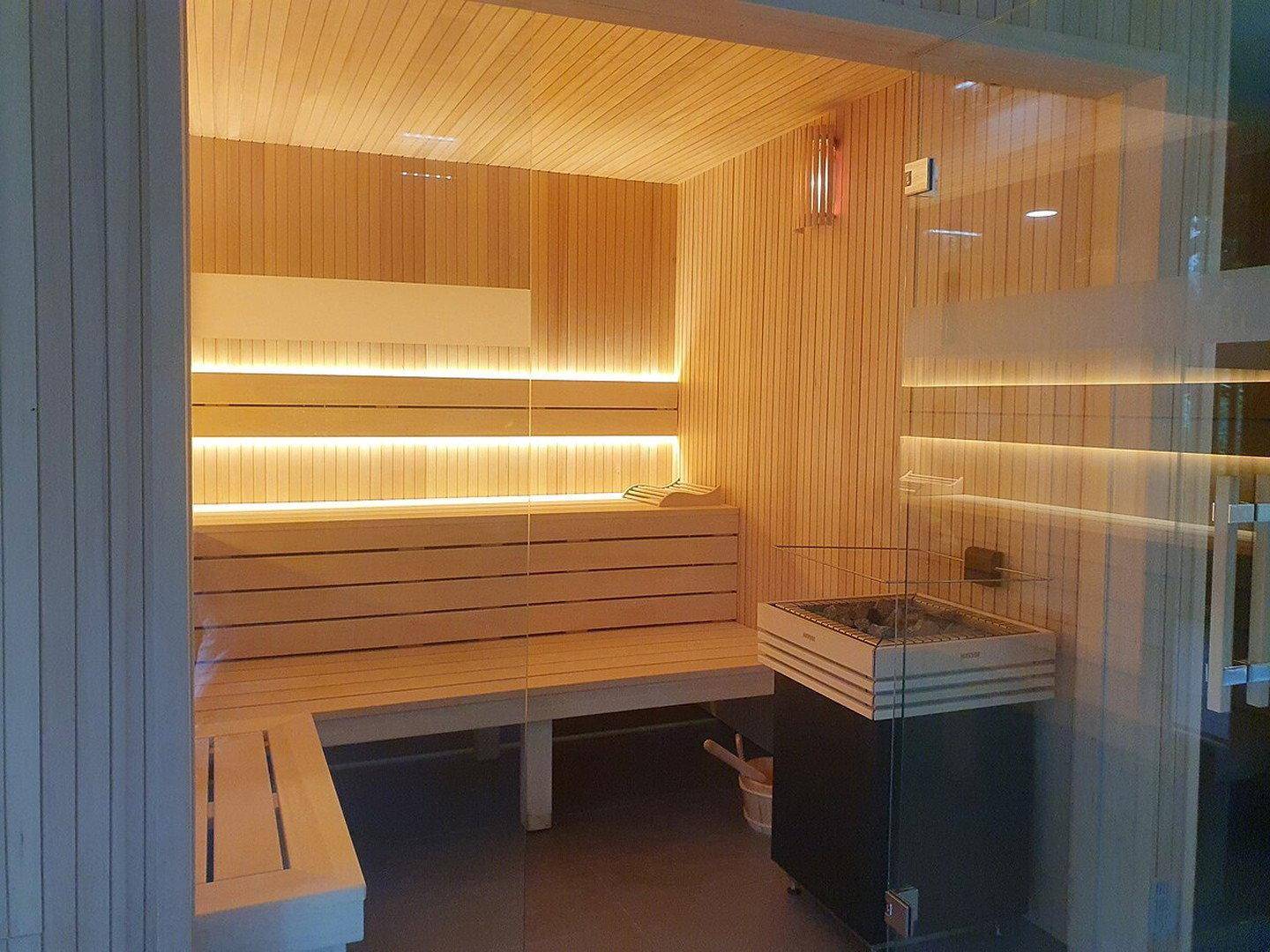 Moderne Sauna mit Holzverkleidung, Sitzbänken und einem Saunaofen hinter einer Glastür.