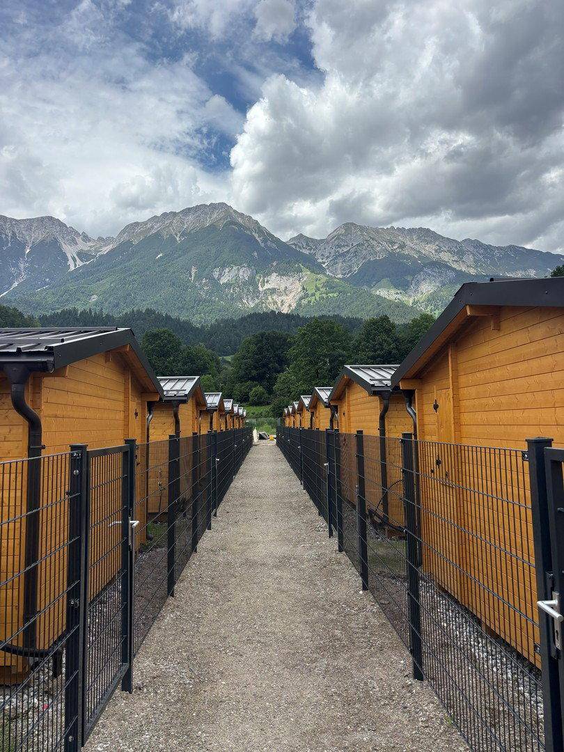 Schmaler Weg zwischen Reihen von Holzhütten mit Zäunen und majestätischen Bergen im Hintergrund.