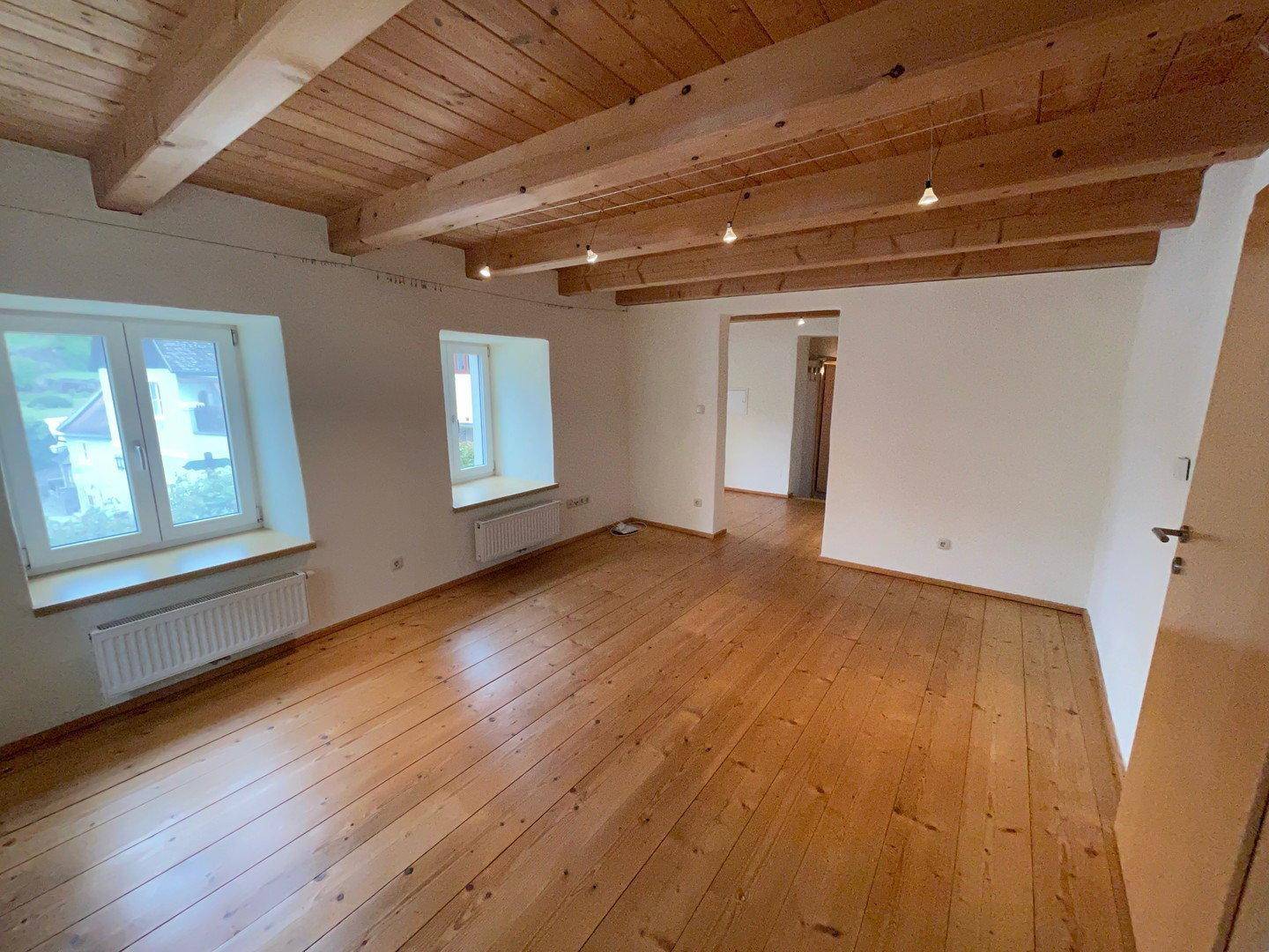 Geräumiges Wohnzimmer mit Holzdielenboden, sichtbaren Deckenbalken und mehreren Fenstern.