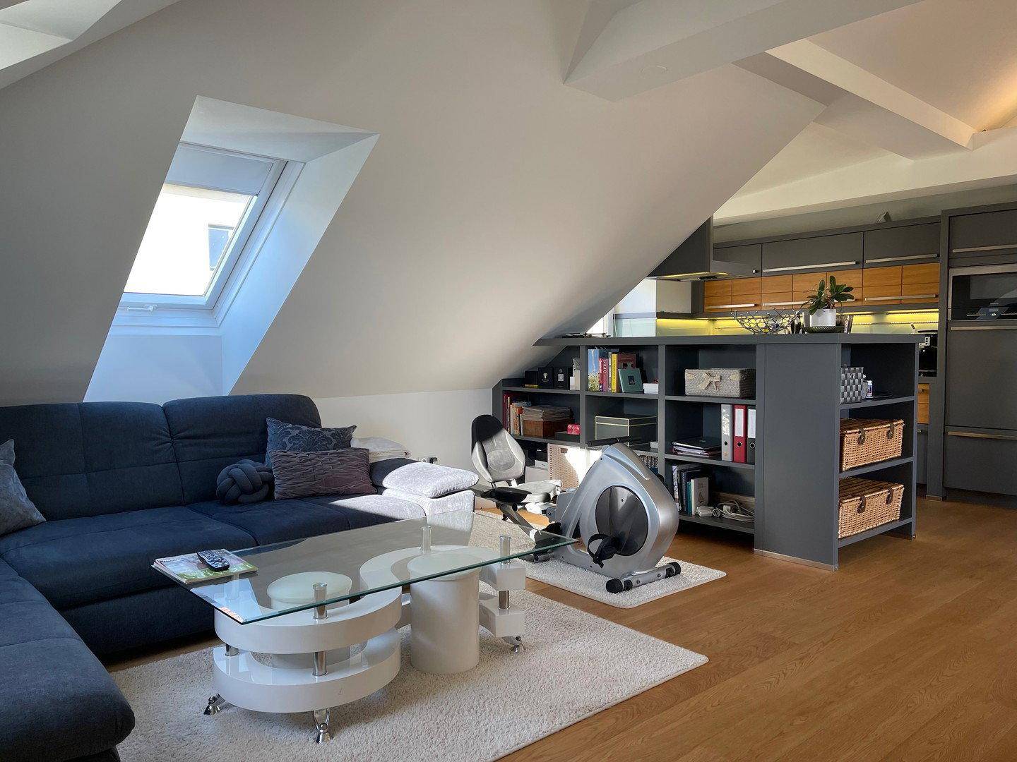 Gemütlicher Wohnbereich mit Dachschrägen, Sofa und einem Heimtrainer, offen zur Küche.