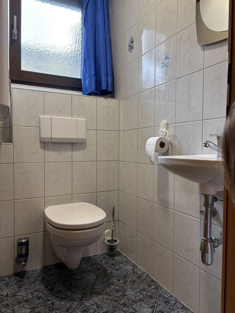 Gästetoilette mit weißen Fliesen, wandhängendem WC, kleinem Waschbecken und einem Fenster mit blauem Vorhang.