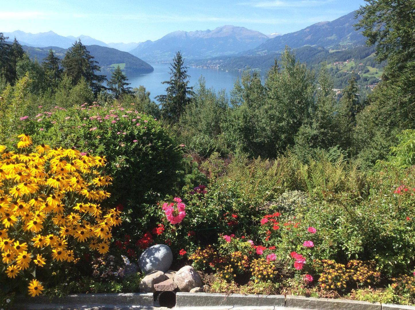 Blühender Garten mit farbenfrohen Pflanzen und einem weiten Blick auf den See und die umliegenden Berge.