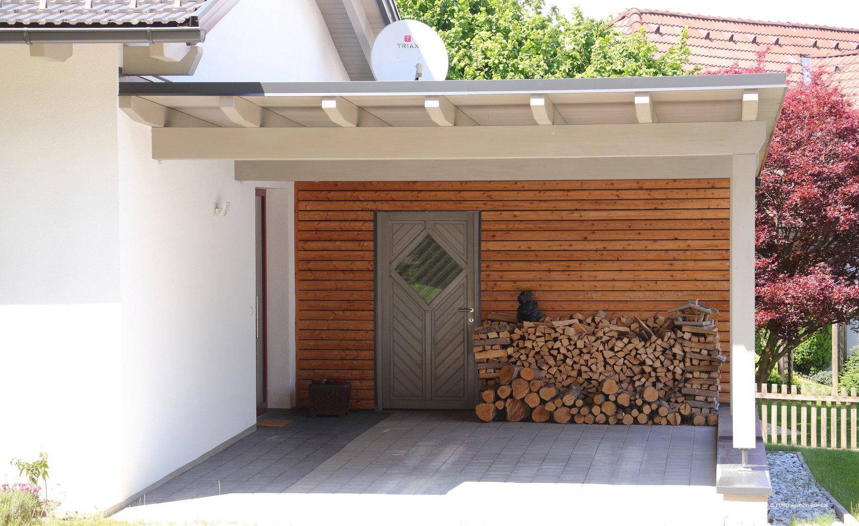 Überdachter Eingangsbereich mit Holzverkleidung und gestapeltem Brennholz, neben einem Carport.