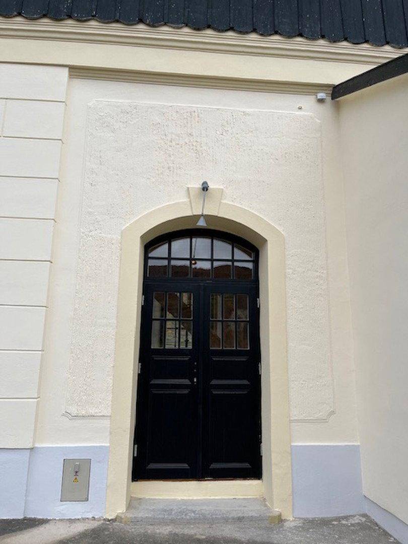 Detailansicht des Haupteingangs mit schwarzer Doppeltür und Bogenfenster in cremefarbener Fassade.