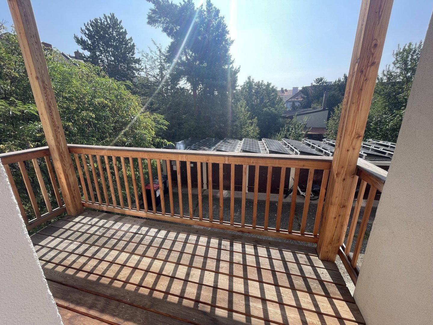 Sonniger Holzbalkon mit Geländer und Blick auf Bäume sowie Solaranlagen im Hintergrund.