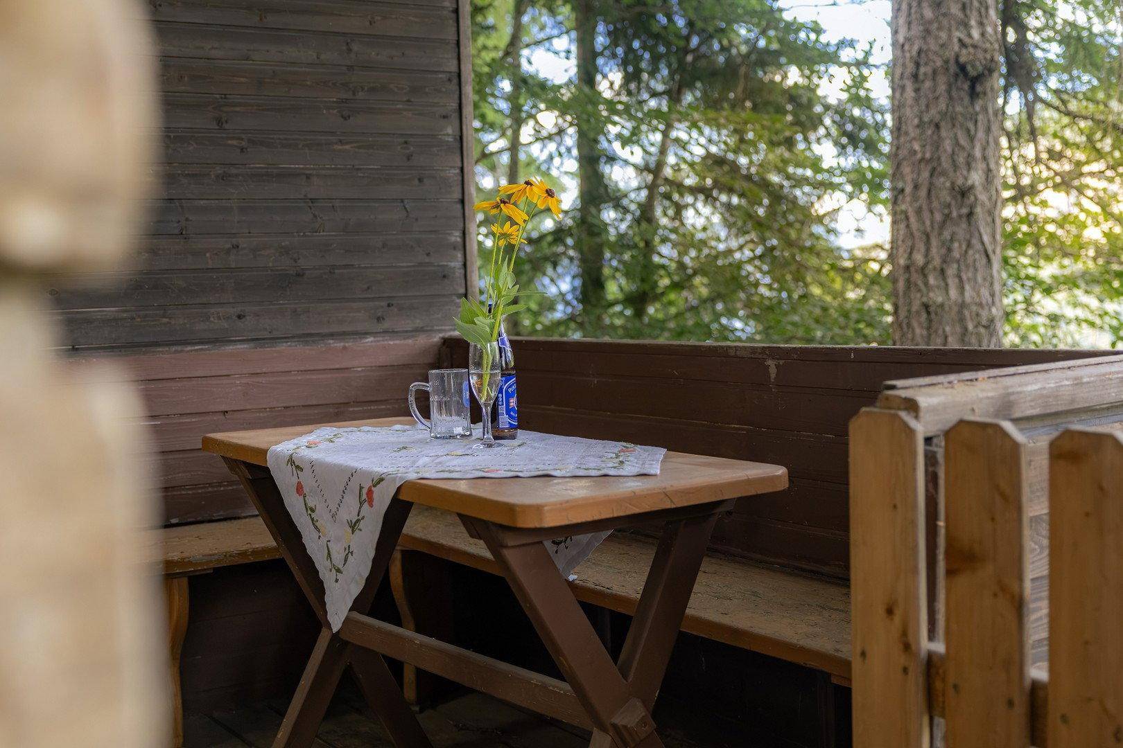 Gemütlicher Außenbereich auf einem Holzbalkon mit Tisch und Bänken, umgeben von Bäumen.