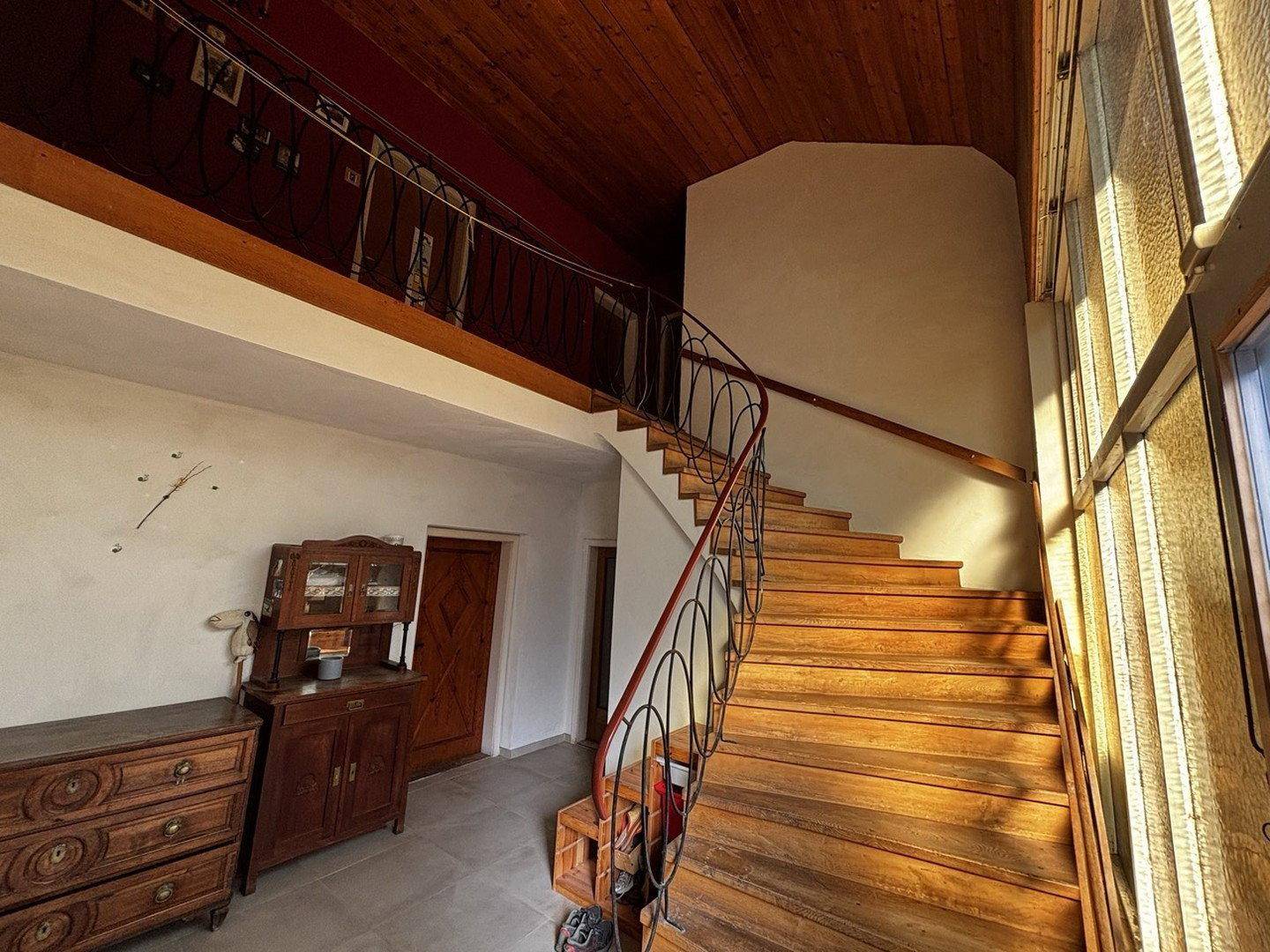 Heller Eingangsbereich mit Holztreppe, schmiedeeisernem Geländer und Holzbalkendecke.