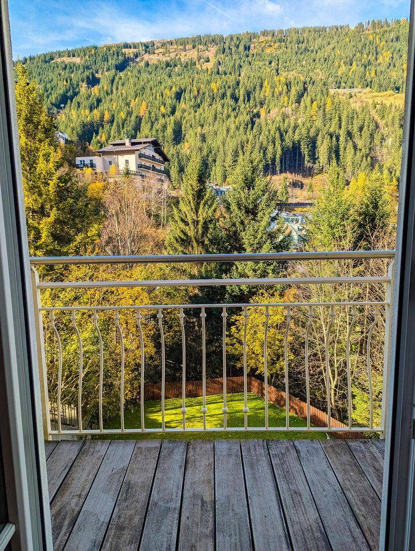 Balkon mit Holzboden und Blick auf die herbstliche Waldlandschaft und ein Gebäude am Hang.
