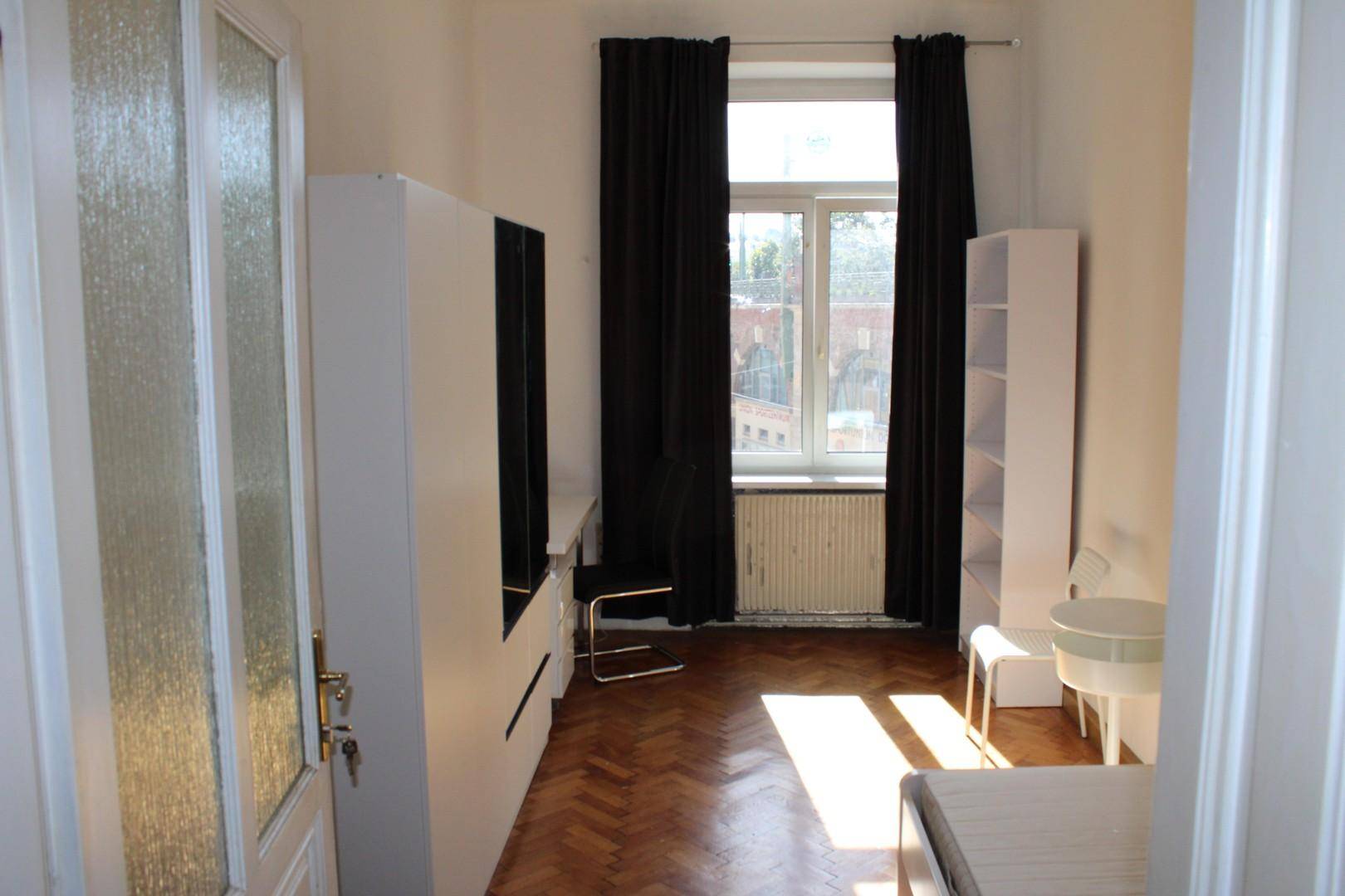 Helles Schlafzimmer mit Parkettboden, weißen Möbeln und einem großen Fenster mit dunklen Vorhängen.