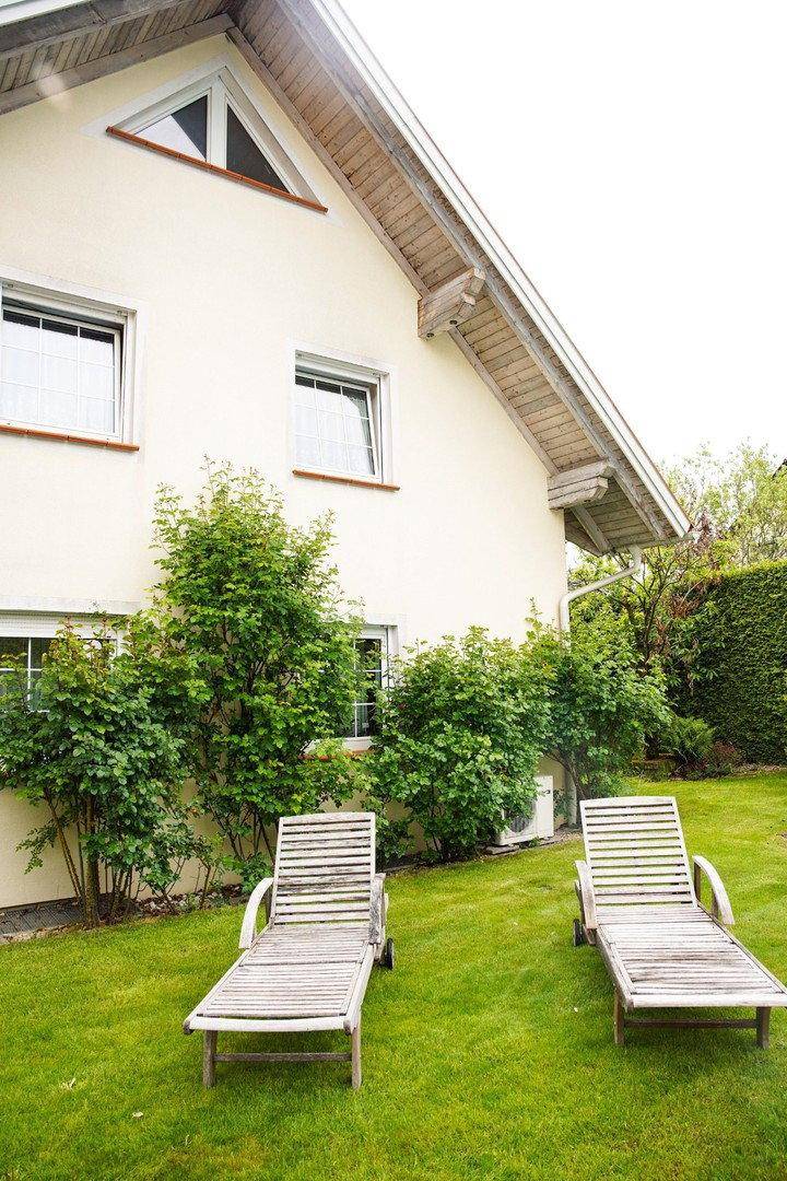 Hausfassade mit Fenstern und zwei Holzliegestühlen auf der grünen Rasenfläche im Garten.