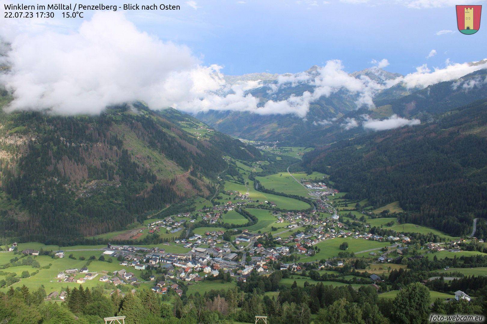 Panoramablick auf ein grünes Tal mit einem Dorf, umgeben von bewaldeten Bergen und Wolken.