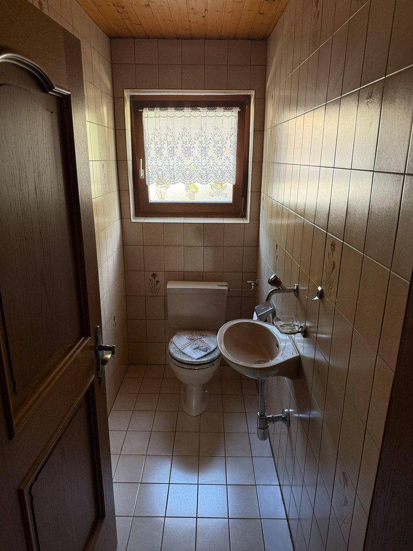 Kleines Gäste-WC mit Fenster, Toilette und Handwaschbecken in beigen Fliesen.