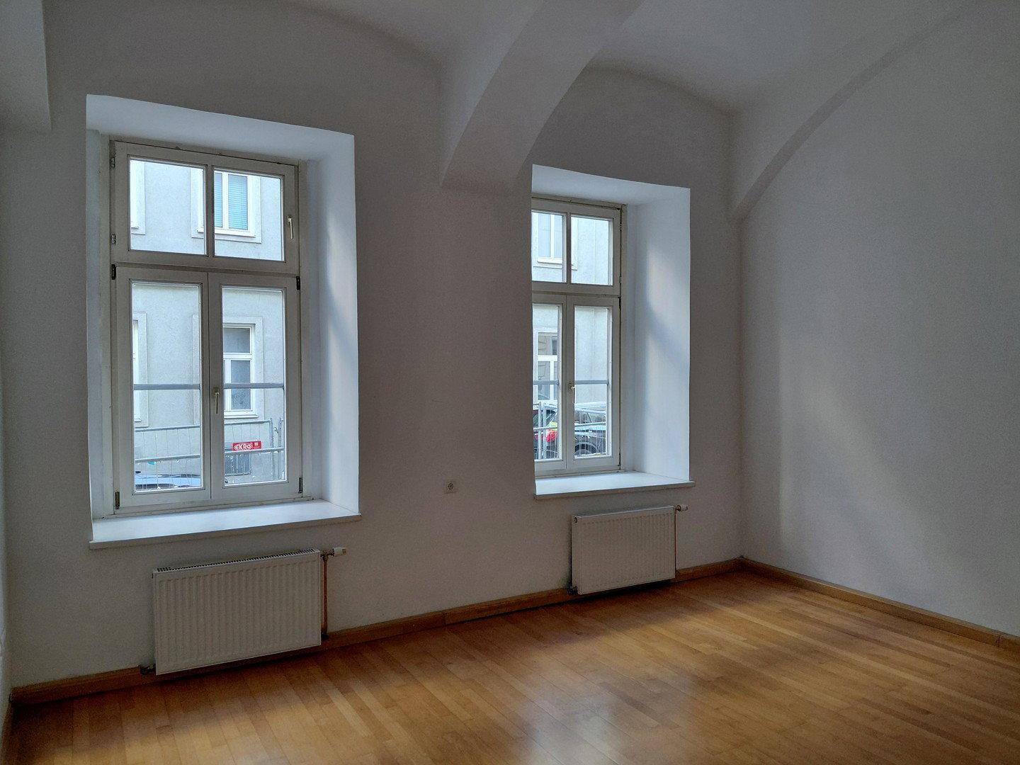 Heller Raum mit zwei großen Fenstern, Holzboden und gewölbter Decke.