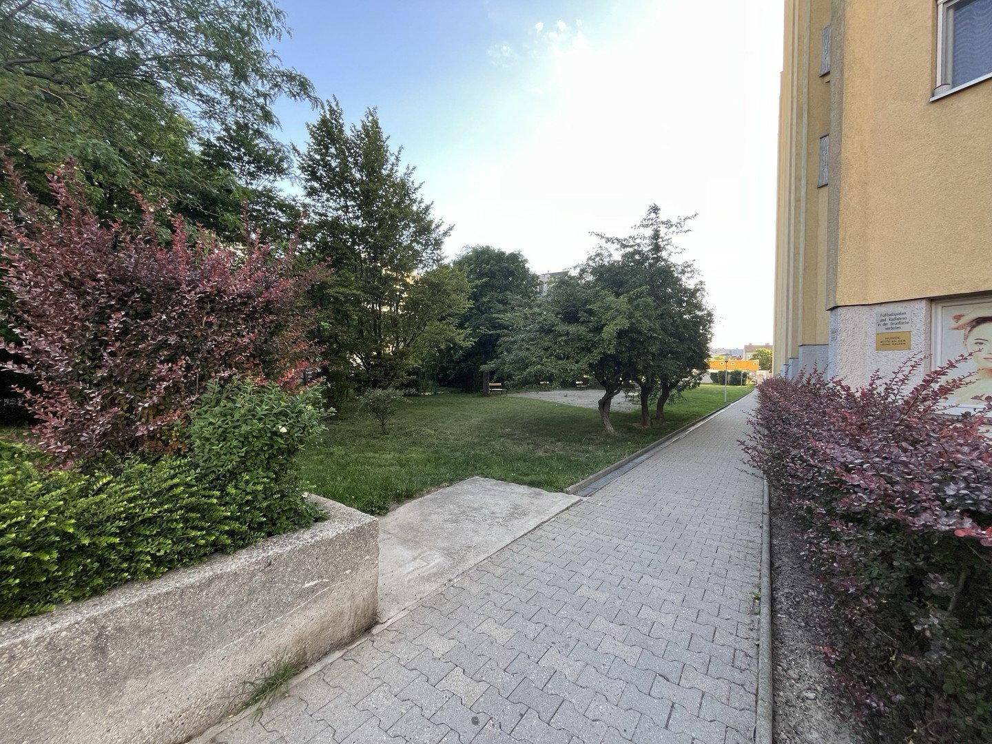 Gepflasterter Weg führt durch eine grüne Gartenanlage mit Büschen und Bäumen neben einem Gebäude.